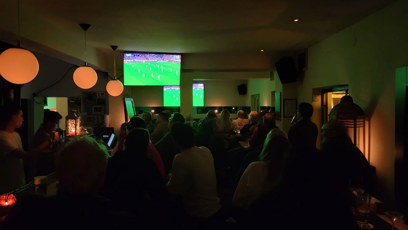 Menschen in einer Bar schauen ein Fußballspiel auf einem Bildschirm an, gedämpftes Licht, entspannte Atmosphäre.